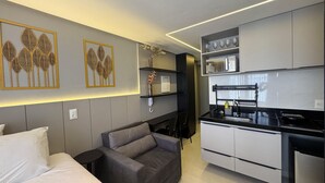 Une cuisine moderne avec des armoires blanches, un comptoir noir et un four encastré. On y trouve un canapé gris avec des coussins et une petite table d'appoint noire. Des œuvres d'art décoratives représentant des motifs de feuilles ornent le mur.