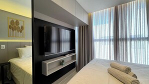 Une chambre d’hôtel moderne dotée d’un grand téléviseur à écran plat fixé au mur, d’un lit avec du linge de lit blanc, d’une table de chevet et d’une petite plante en pot.