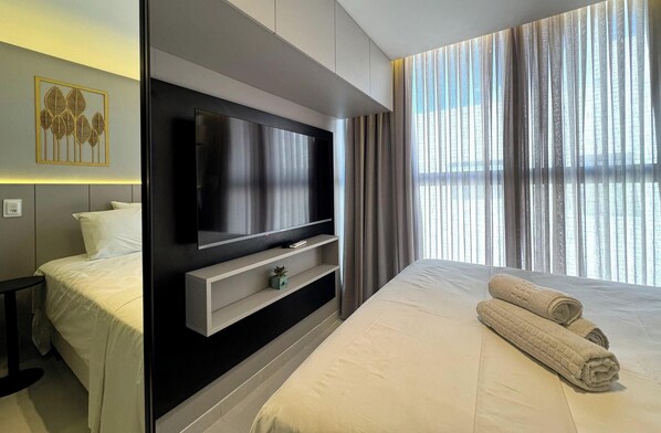 Une chambre d’hôtel moderne dotée d’un grand téléviseur à écran plat fixé au mur, d’un lit avec du linge de lit blanc, d’une table de chevet et d’une petite plante en pot.