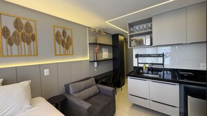 A modern kitchen with white cabinets, a black countertop, and a built-in oven. There is a grey sofa with cushions and a small black side table. Decorative wall art with leaf patterns is visible. - Breeze Miramar Hospedagens (Joao Pessoa)