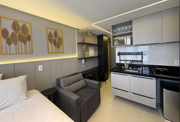 Una cocina moderna con armarios blancos, encimera negra y horno empotrado. Hay un sofá gris con cojines y una pequeña mesita auxiliar negra. Se observa arte decorativo en la pared con motivos de hojas.