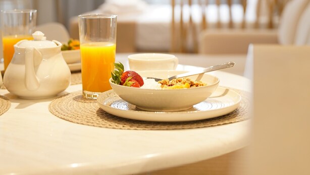 A breakfast table with a teapot, glasses of orange juice, a bowl of cereal with a strawberry, and a spoon.