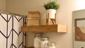 A bathroom with a shower curtain, a wooden shelf with jars, a potted plant, and a framed picture on the wall. - Local Gem w/ King, 65” TV, Hot Tub, Ski Bus (Park City)