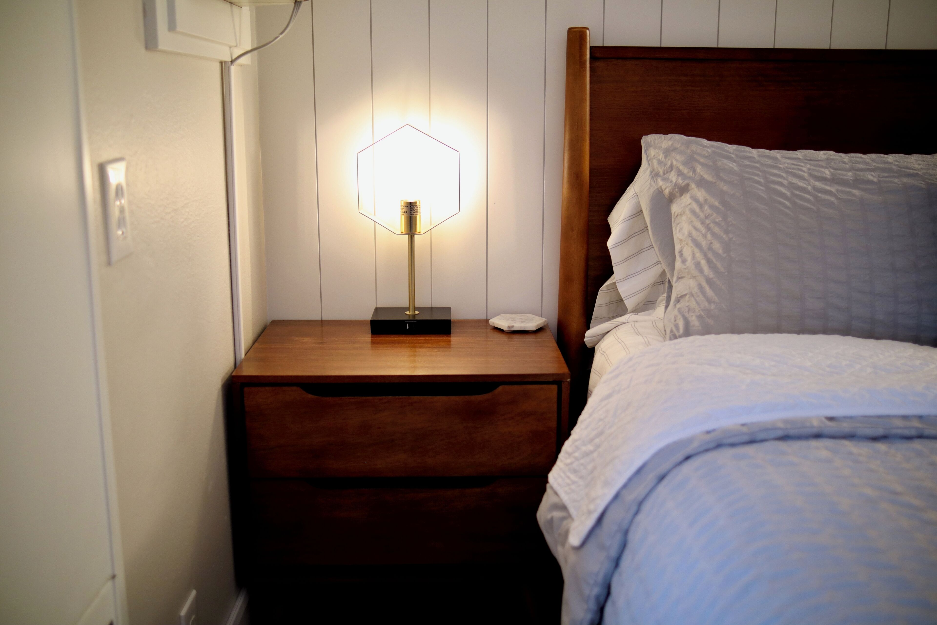 Un dormitorio con cabecera de madera, una mesita de noche con lámpara y una cama con ropa blanca.