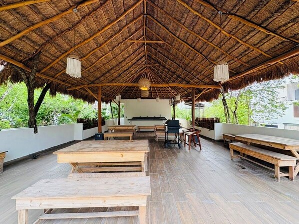 An open-air dining area with wooden benches and tables, a thatched roof, and a view of greenery outside.
