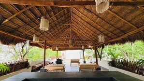A covered outdoor seating area with wooden benches and a central fire pit, surrounded by lush greenery. - Beautiful apartment 10 minutes from the beach with Jacuzzi Swimming pools GYM Terraces. (TULUM)