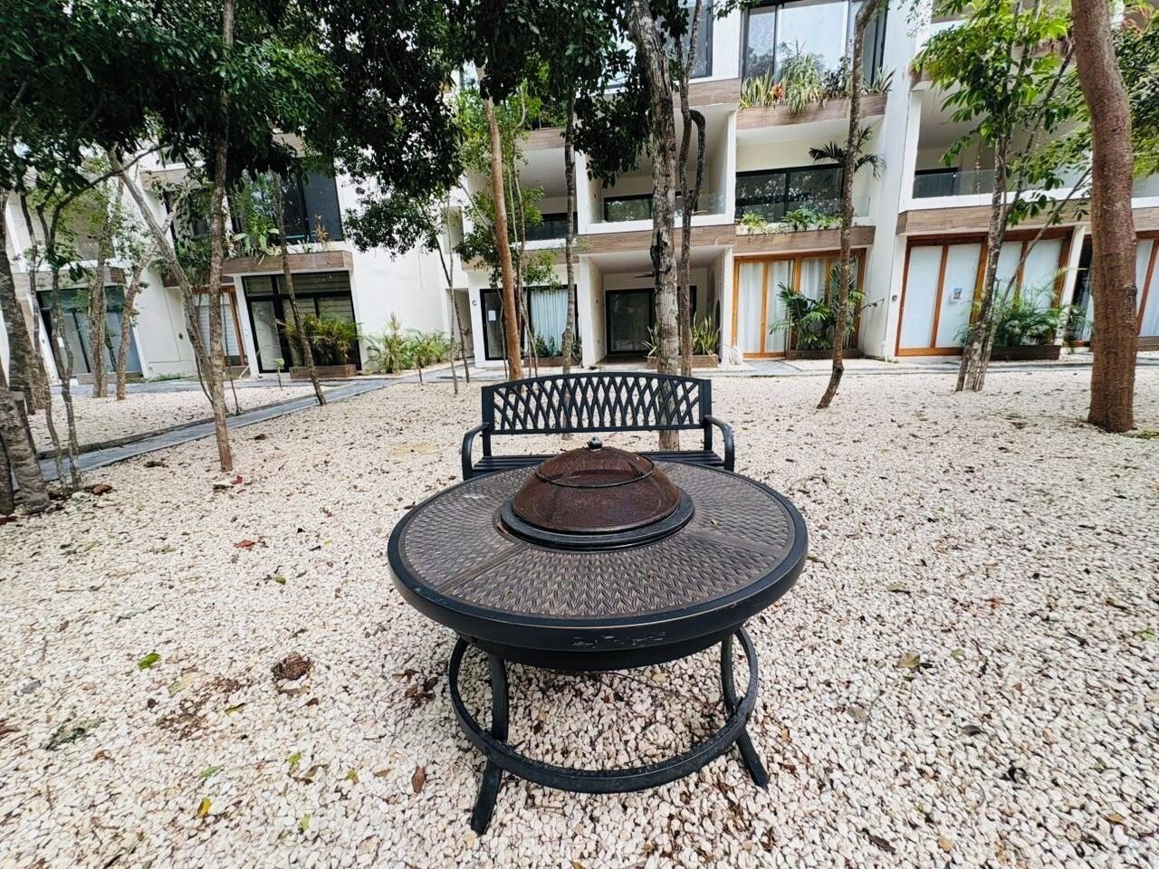 A round outdoor table with a metal frame and a woven tablecloth, surrounded by a gravel area with trees and a building in the background.