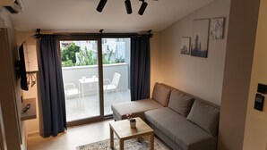 Een woonkamer met een bank, een kleine tafel met een plant, een schuifpui naar een balkon en uitzicht op groen.