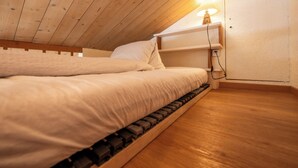 Une chambre avec un plafond en bois, un lit avec une literie blanche, une table de chevet avec une lampe et une étagère fixée au mur.