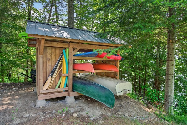 A wooden structure with a metal roof, surrounded by trees, containing several kayaks and a paddleboard. - Spa & Waterfront | Chalet le Colibri (Amherst)