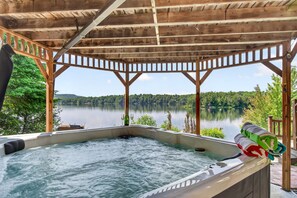 A hot tub with a wooden pergola structure, overlooking a lake with trees.