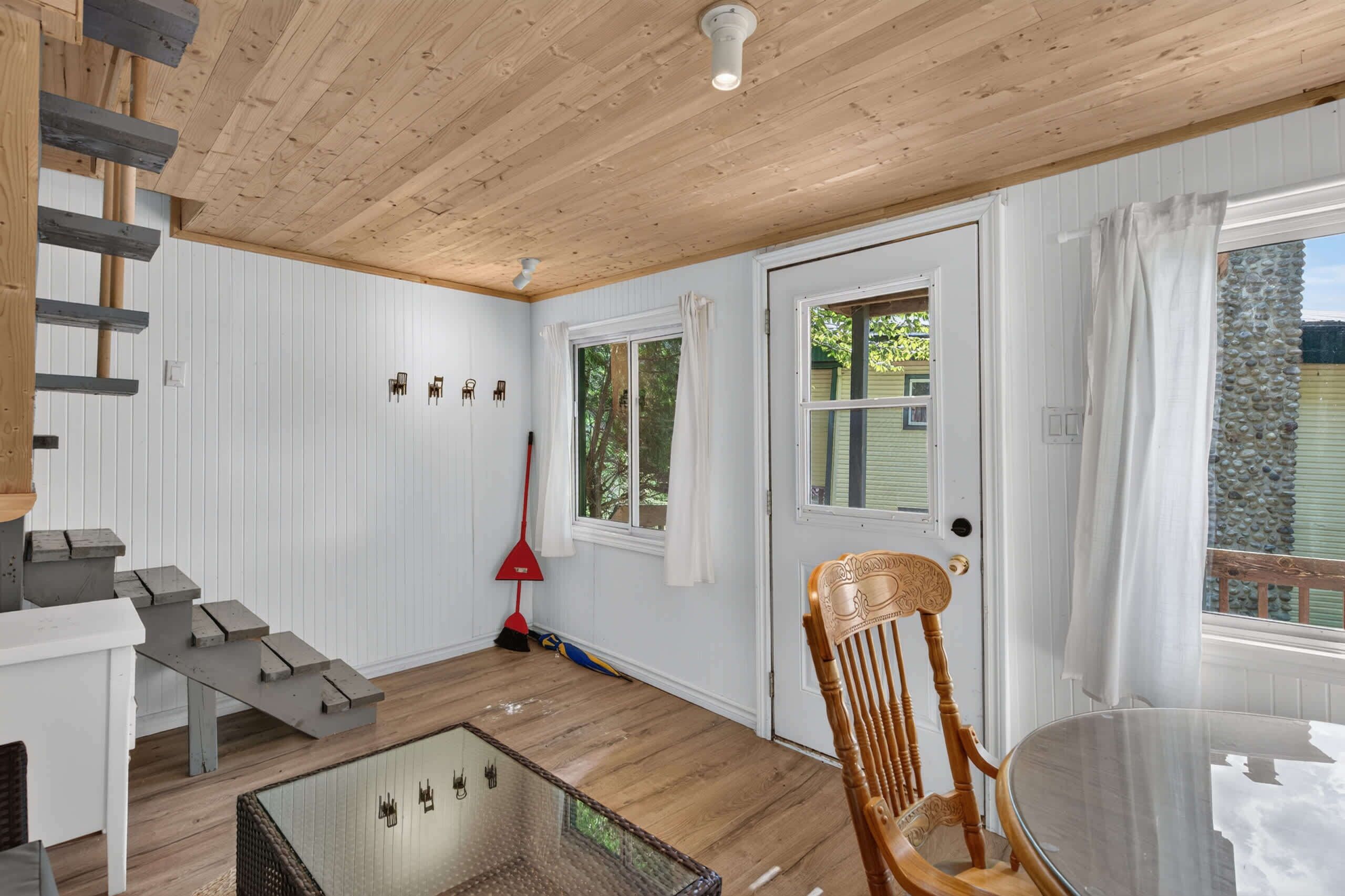 Quarto com teto de madeira, escada, mesa de vidro, cadeira de madeira, porta com janela e uma vassoura vermelha.