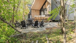 Una cabaña moderna con terraza de madera, mobiliario exterior y un amplio ventanal con vistas a un área boscosa.