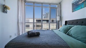 Un dormitorio con un ventanal grande, cama con ropa de cama verde y gris, y vista a la ciudad.