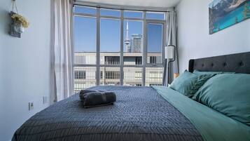 A bedroom with a large window, a bed with green and grey bedding, and a cityscape view.