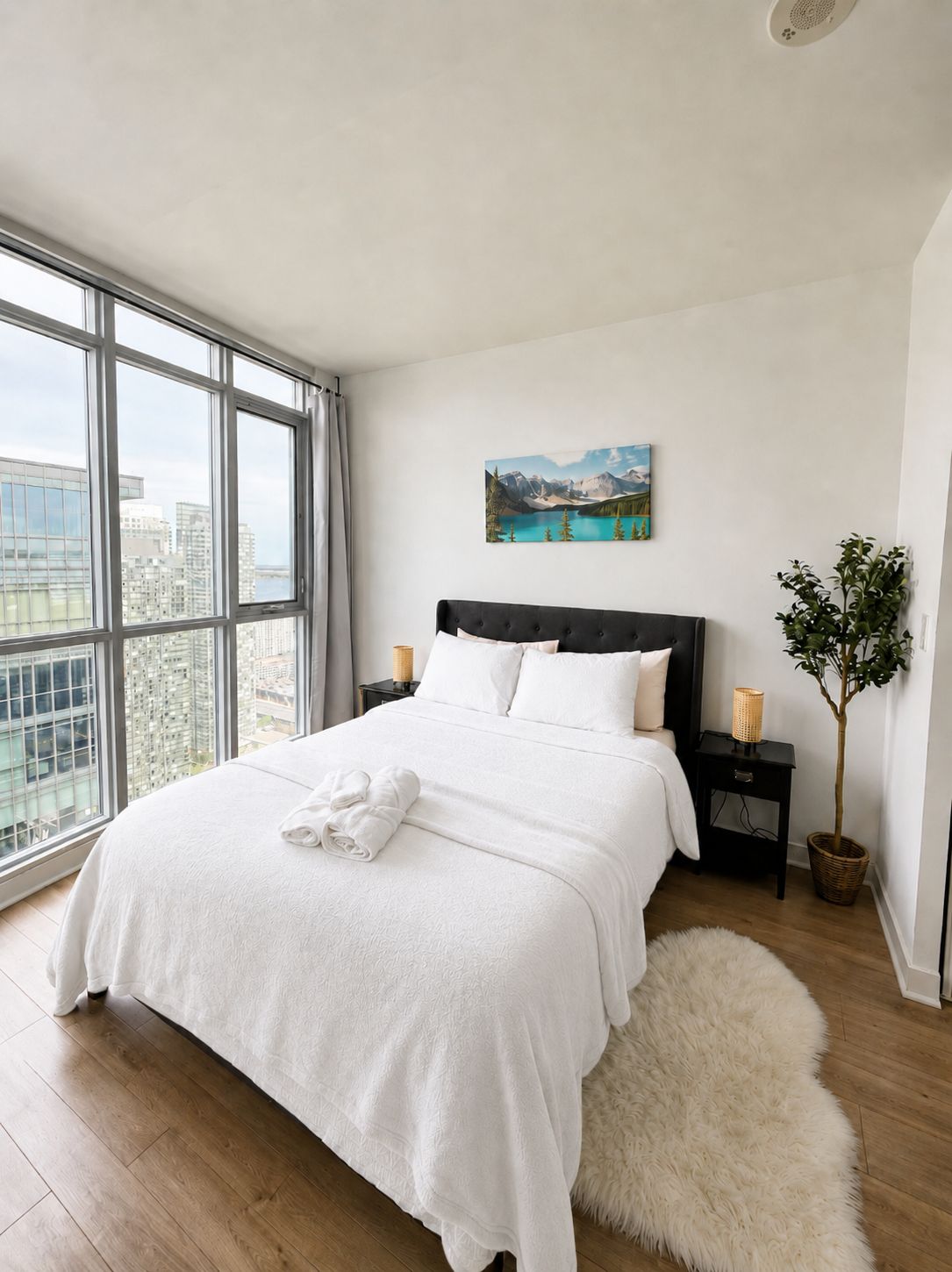 A modern bedroom with a large window, a bed with white bedding, a painting, and a small plant.