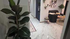 Un salon moderne avec un canapé noir, une table basse et un coin repas comprenant une table et des chaises en bois. On aperçoit, au premier plan, une grande plante en pot et une petite table sur laquelle se trouvent une bouteille et un verre. Les murs de la pièce sont blancs et le sol est recouvert de marbre.