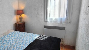 A bedroom with a bed, a bedside table with a lamp, a radiator, and a window with curtains. - Nice 2P with Wifi & swimming pool near city center (Fréjus)