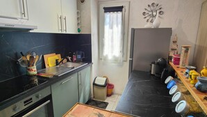 A compact kitchen with a black countertop, a stove, and a sink. There is a window with a curtain, a trash bin, and a shelf with kitchen utensils. - Nice 2P with Wifi & swimming pool near city center (Fréjus)