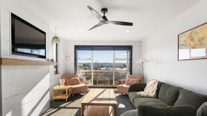 A modern living room with a large window, a ceiling fan, and a flat-screen TV mounted on the wall. - Sunsets, space and family fun- mountain views, trampoline and child`s play area, fully fenced (Taupō)
