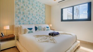 Ein ordentlich bezogenes Bett mit weißer Decke und einem zusammengerollten Handtuch, eine Nachttischlampe und eine Wand mit geometrischem Muster.