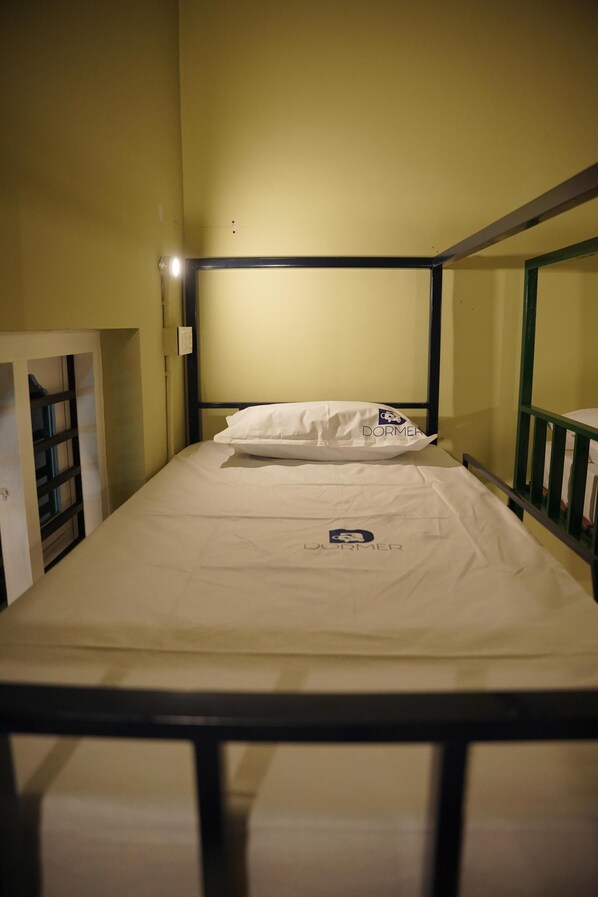 Una cama individual con colchón y almohada blancos, estructura metálica y un pequeño logo en el colchón.