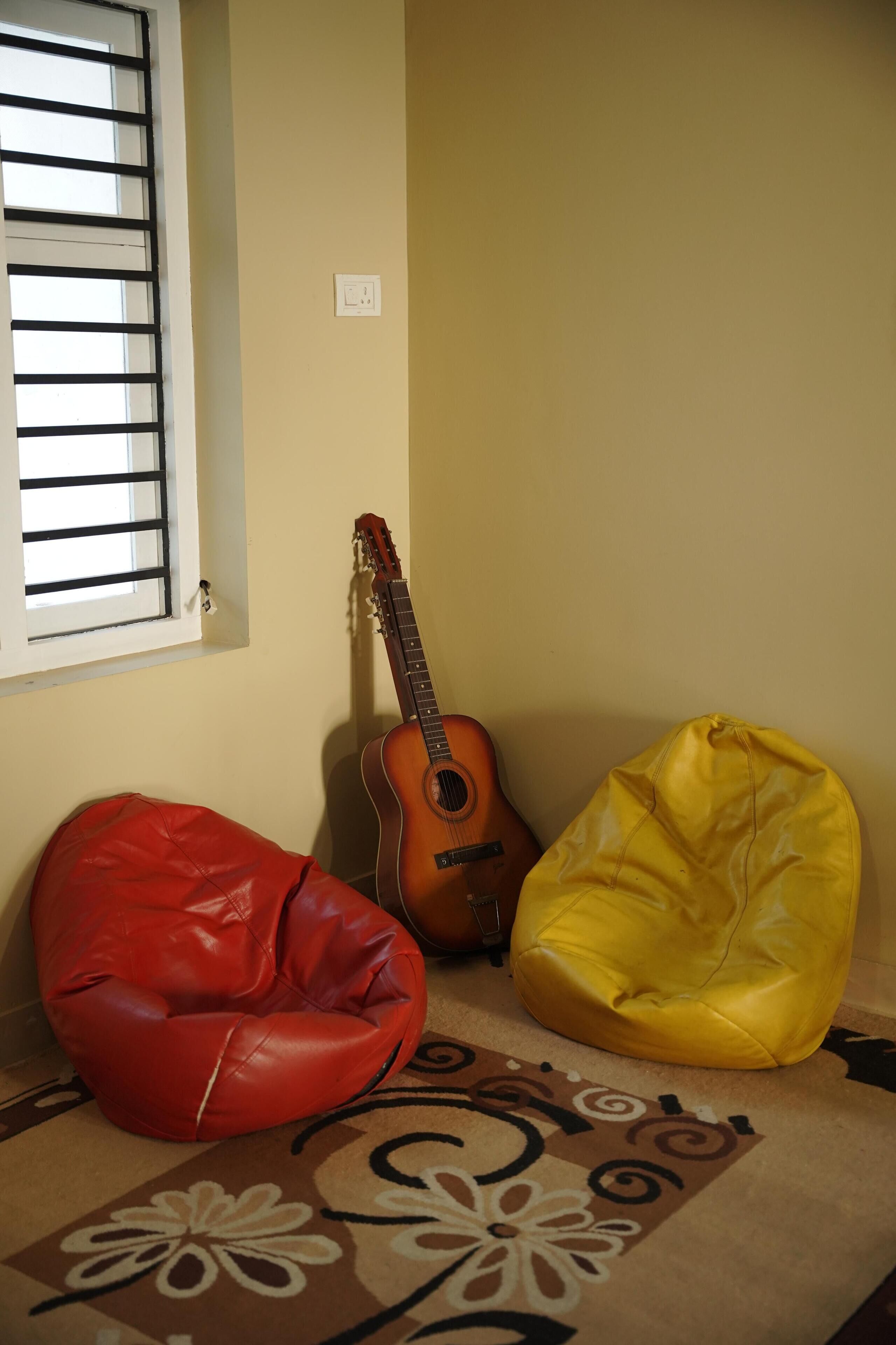 A room with a red and yellow bean bag chair, a guitar, and a window with blinds.
