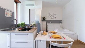 A modern kitchen with a sink, stove, and countertop. There is a dining table with plates, a loaf of bread, and a vase with flowers. A bed is visible in the background with a wall decoration.