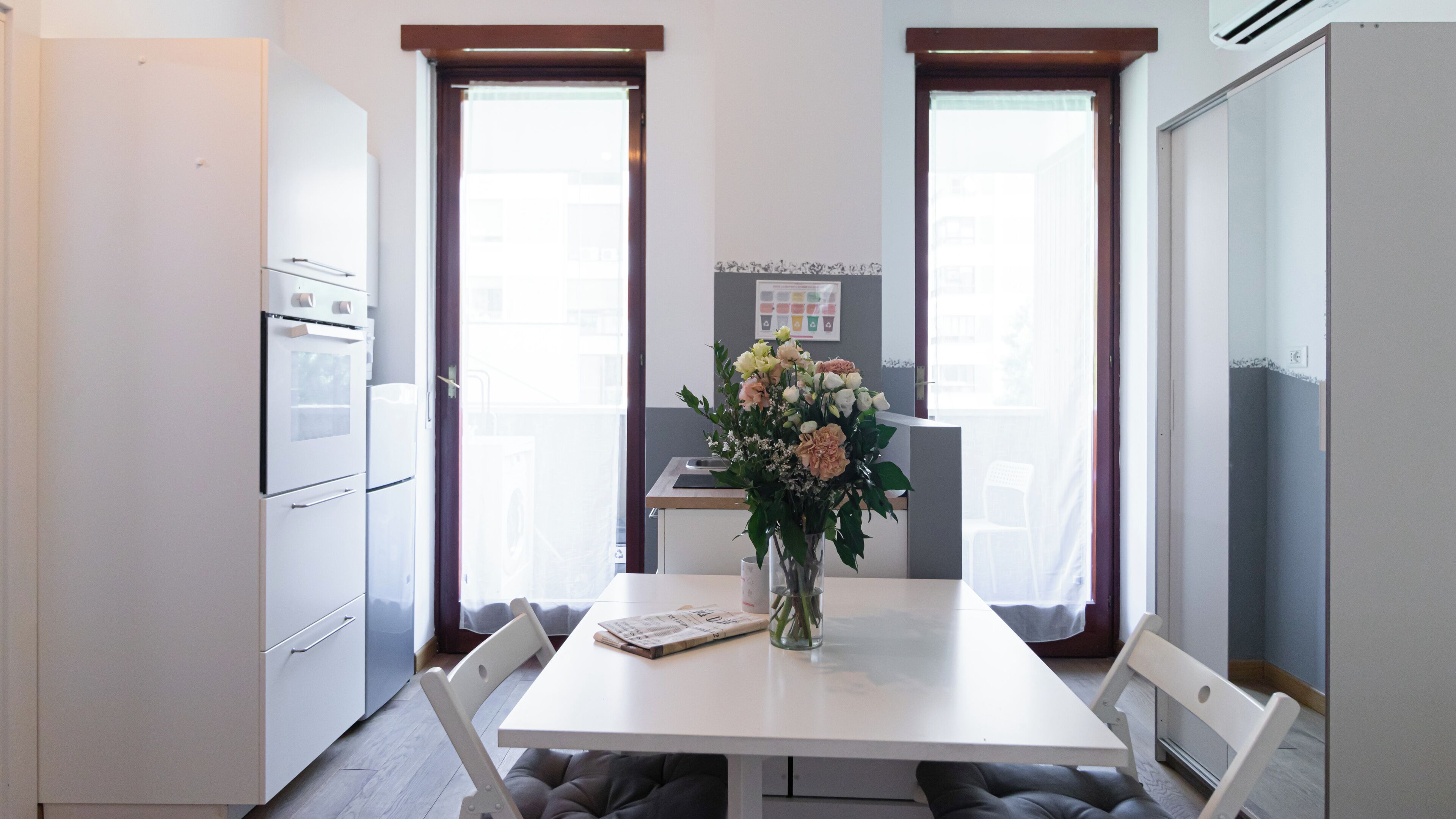 Eine kleine Küche mit weißen Geräten, einem Esstisch mit Blumenvase und Blick durch die Fenster.