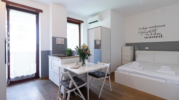 A compact hotel room with a kitchenette, a dining area with a table and chairs, a bed with neatly folded towels, and a motivational quote on the wall.