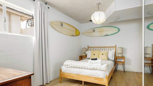 Une chambre avec un lit en bois, du linge de lit blanc, deux planches de surf accrochées au mur et une table de chevet en bois avec une lampe.