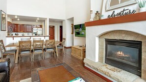 A spacious living room with a fireplace, wooden furniture, and a kitchen in the background.