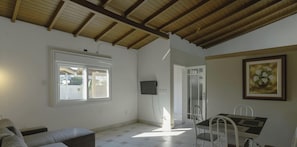 A room with a wooden ceiling, a white couch, a dining table with chairs, a TV mounted on the wall, and a door leading outside.