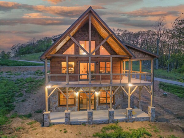 A two-story wooden cabin with a stone foundation, large windows, and a balcony. - Brand New | Hot Tub + Grandfather Mtn + Views (Banner Elk)