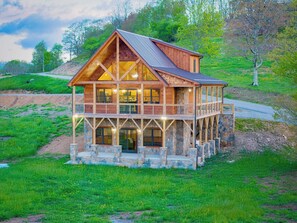A two-story log cabin with a stone foundation, large windows, and a balcony. - Brand New | Hot Tub + Grandfather Mtn + Views (Banner Elk)