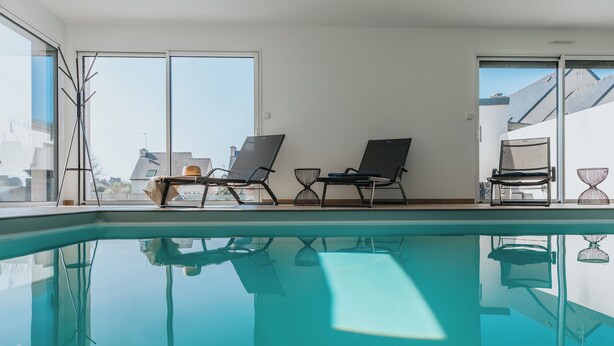 Une piscine intérieure avec des parois en verre transparent, deux chaises longues et une petite table avec une chaise.