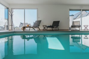 An indoor swimming pool with clear glass walls, two lounge chairs, and a small table with a chair.