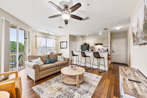 A modern living room with a sofa, coffee table, and chairs, a kitchen area with bar stools, and a ceiling fan.