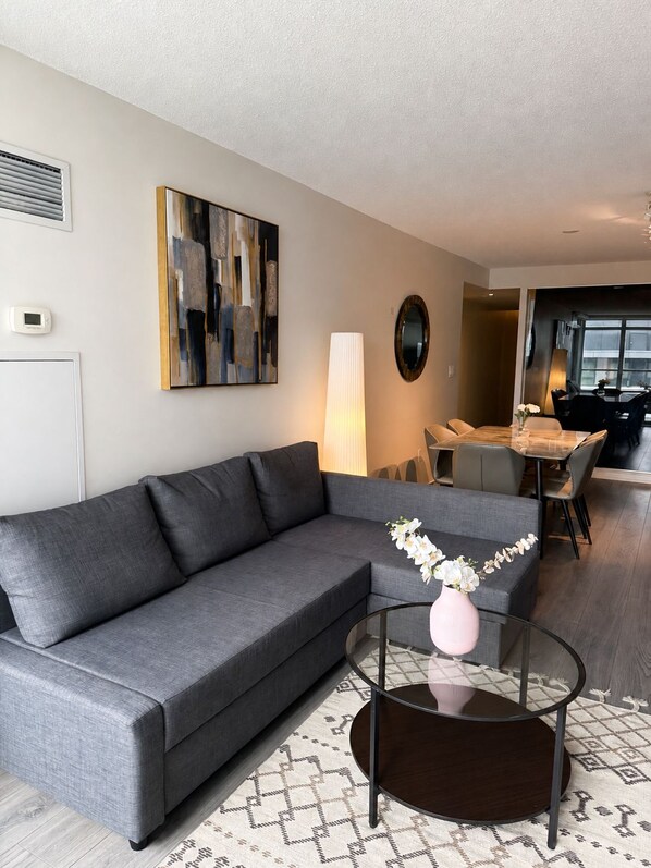 A modern living room with a grey sofa, a glass coffee table with a pink vase, a painting on the wall, and a dining area in the background.
