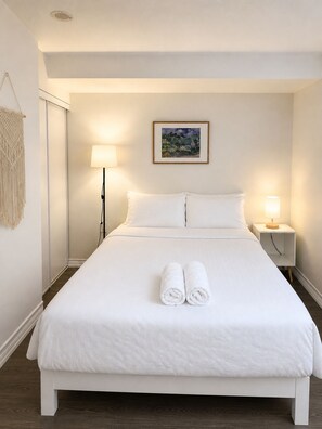 Een net opgemaakt bed met witte lakens en twee opgerolde handdoeken, een bedlamp en een ingelijste foto aan de muur.