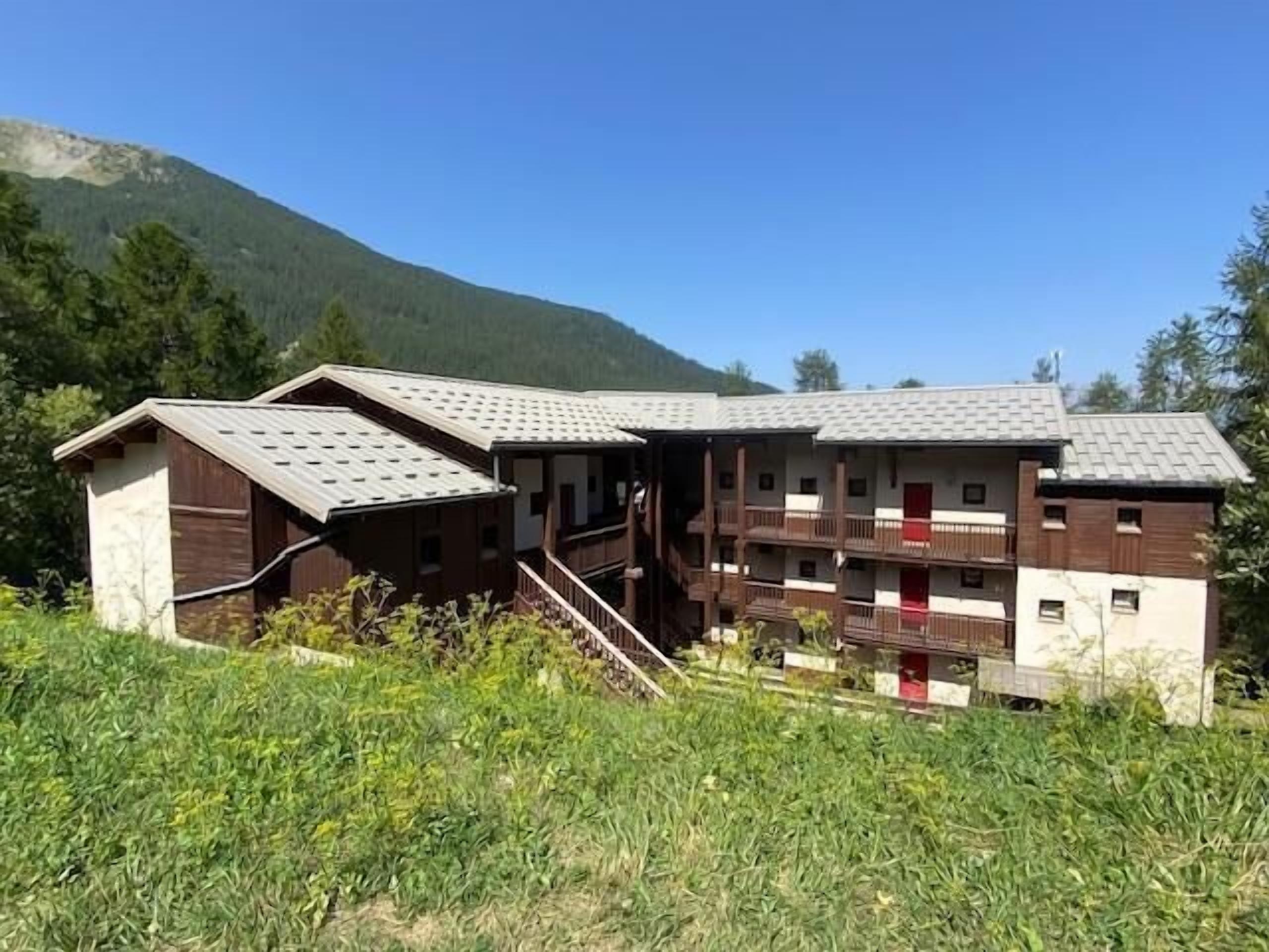 A two-story building with a wooden exterior and a red door, surrounded by greenery and mountains.