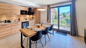 A modern kitchen with wooden cabinets, a dining table set for four, and a view of a mountainous landscape through large sliding glass doors. - Cumaru-appartement-4pers-Proche Centre (Les Gets)