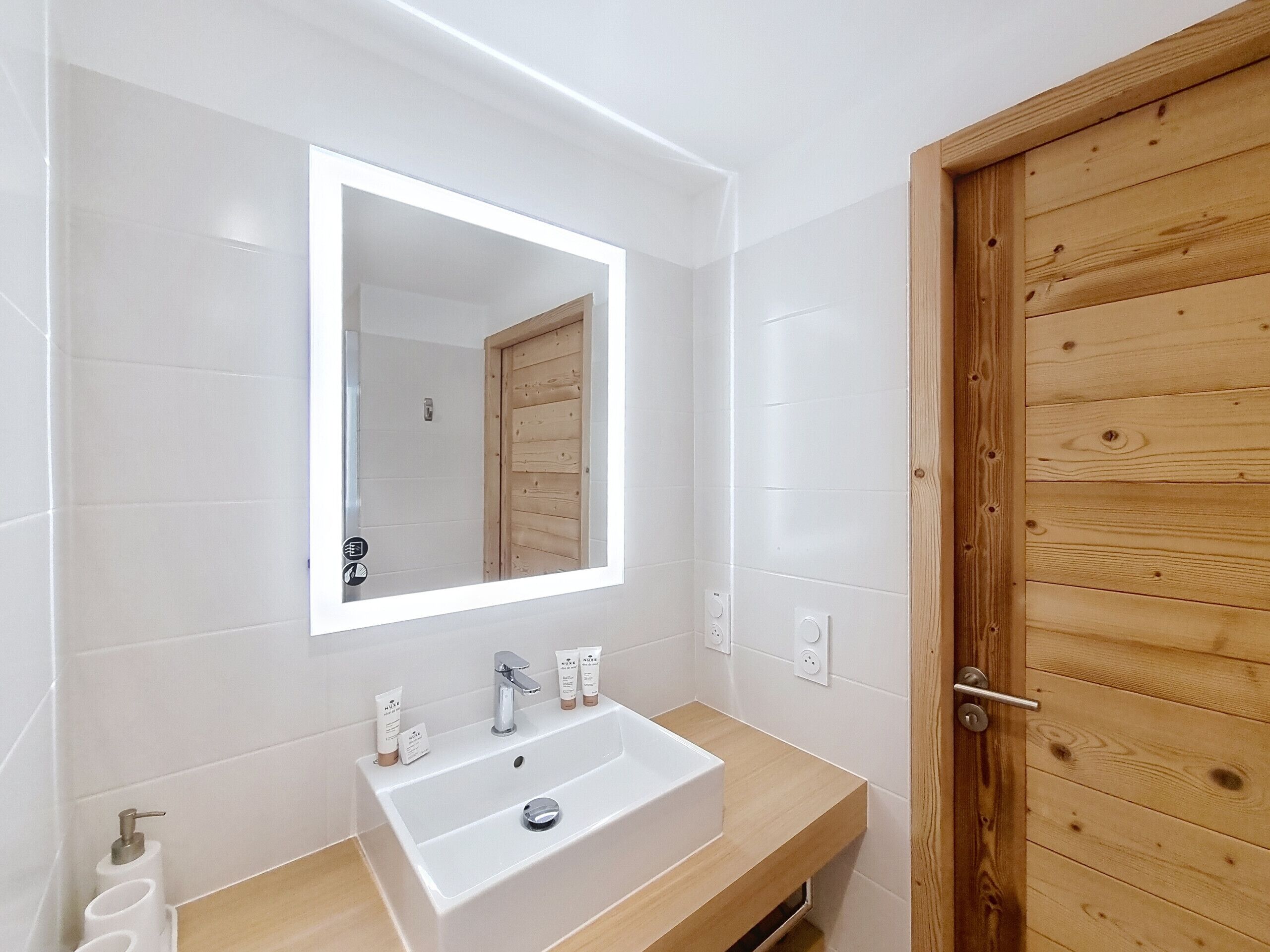 A modern bathroom with a white sink, a large mirror, and wooden door.