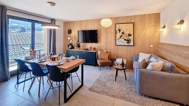 A modern living room with a dining area, wooden panel walls, a flat-screen TV, and a large window offering a view of the roof. - Cumaru-appartement-4pers-Proche Centre (Les Gets)