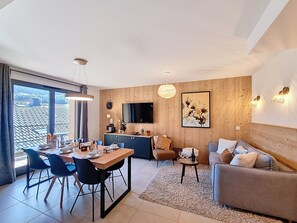 Un salon moderne comprenant un coin repas, des murs recouverts de panneaux en bois, un téléviseur à écran plat et une grande fenêtre donnant sur le toit.