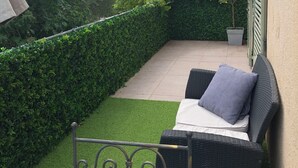 Un patio con un sofá de mimbre y cojines, un seto bien cuidado y vistas a un edificio con tejado de tejas.