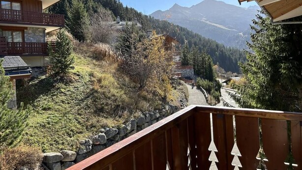 Vista sulle montagne da un balcone con ringhiera in legno, che si affaccia su una valle con case e alberi.