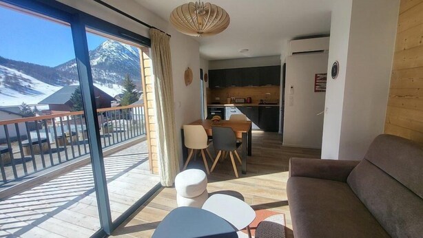 Una sala de estar moderna con un sofá, una mesa de comedor con sillas, una zona de cocina y un balcón con vistas a las montañas.