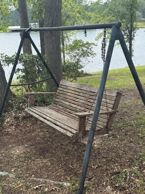 A wooden bench with metal chains, set on a dirt ground with trees and a body of water in the background.