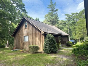 A rustic wooden barn with a sloped roof, surrounded by lush greenery and trees.